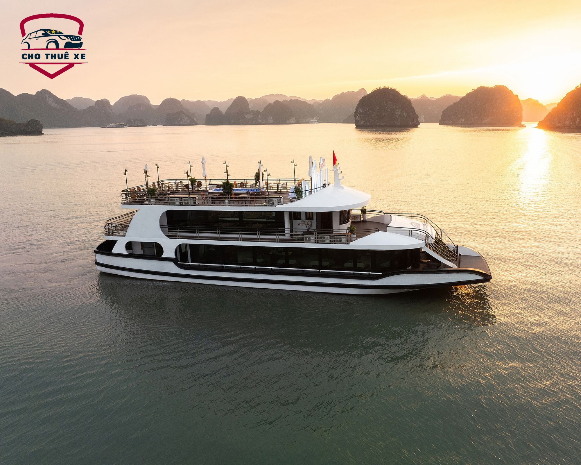 Du thuyền hiện đại với nhiều tầng và boong tắm nắng, đang tham quan Vịnh Hạ Long trong ánh nắng chiều tà, bao quanh là các đảo đá vôi đặc trưng