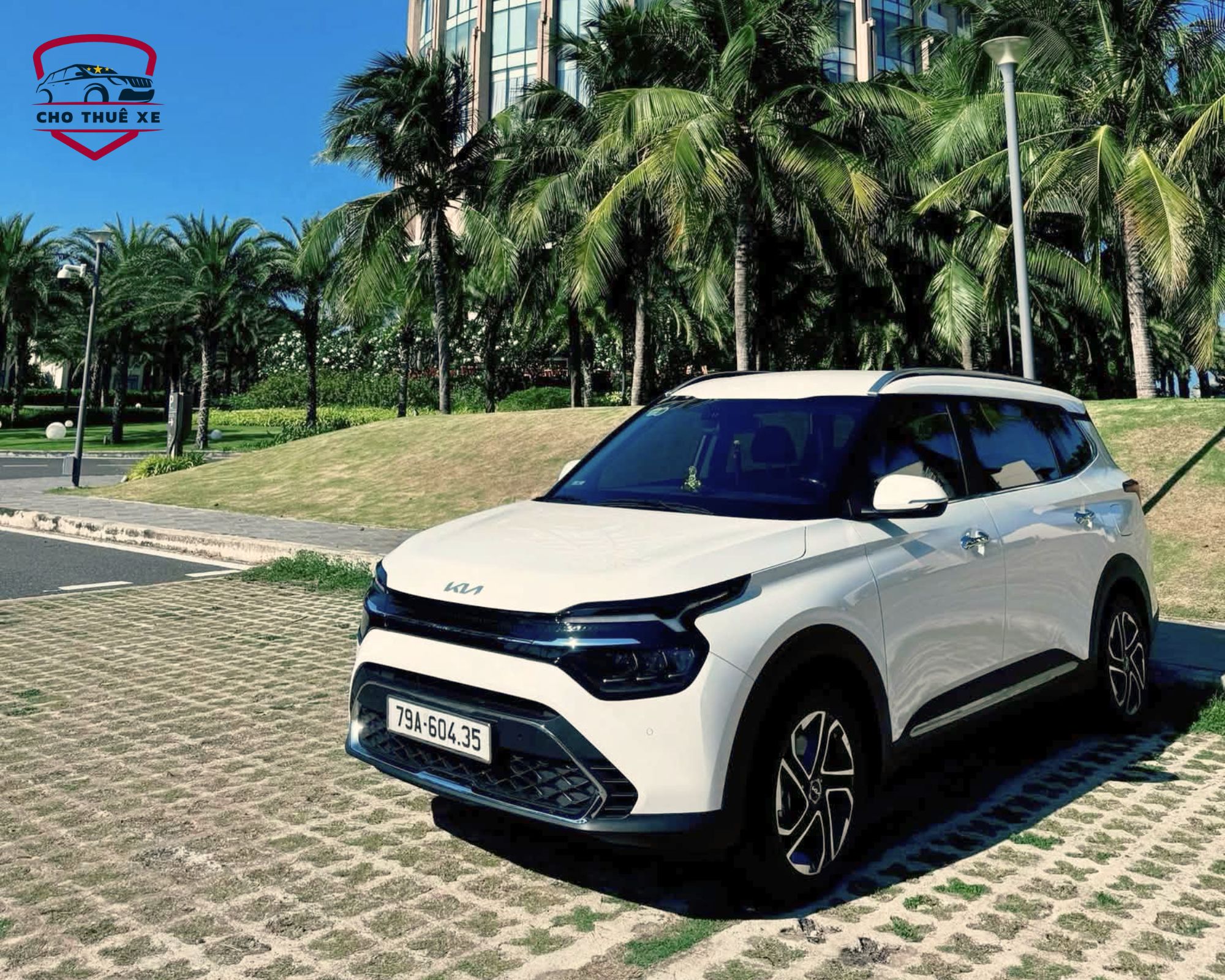 Xe Kia Carens SUV trắng sạch sẽ đậu trước khuôn viên cây dừa xanh mát, dịch vụ thuê xe riêng tiện lợi cho chuyến du lịch Nha Trang